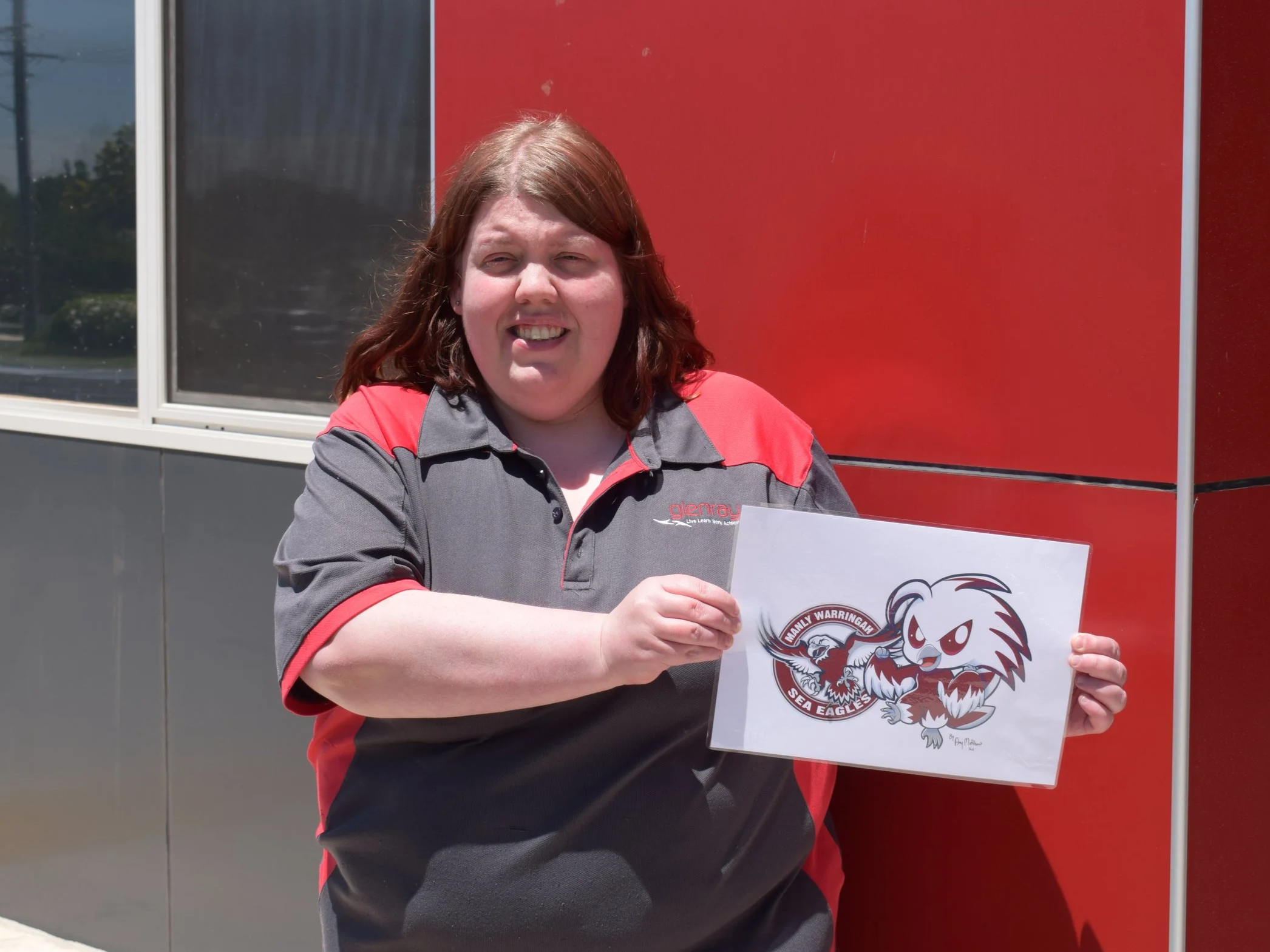 Young woman wearing a Glenray branded polo. She is holding up her artwork and smiling. The artwork features the Manly Sea Eagles logo next to a stylised caricature of the eagle mascot.