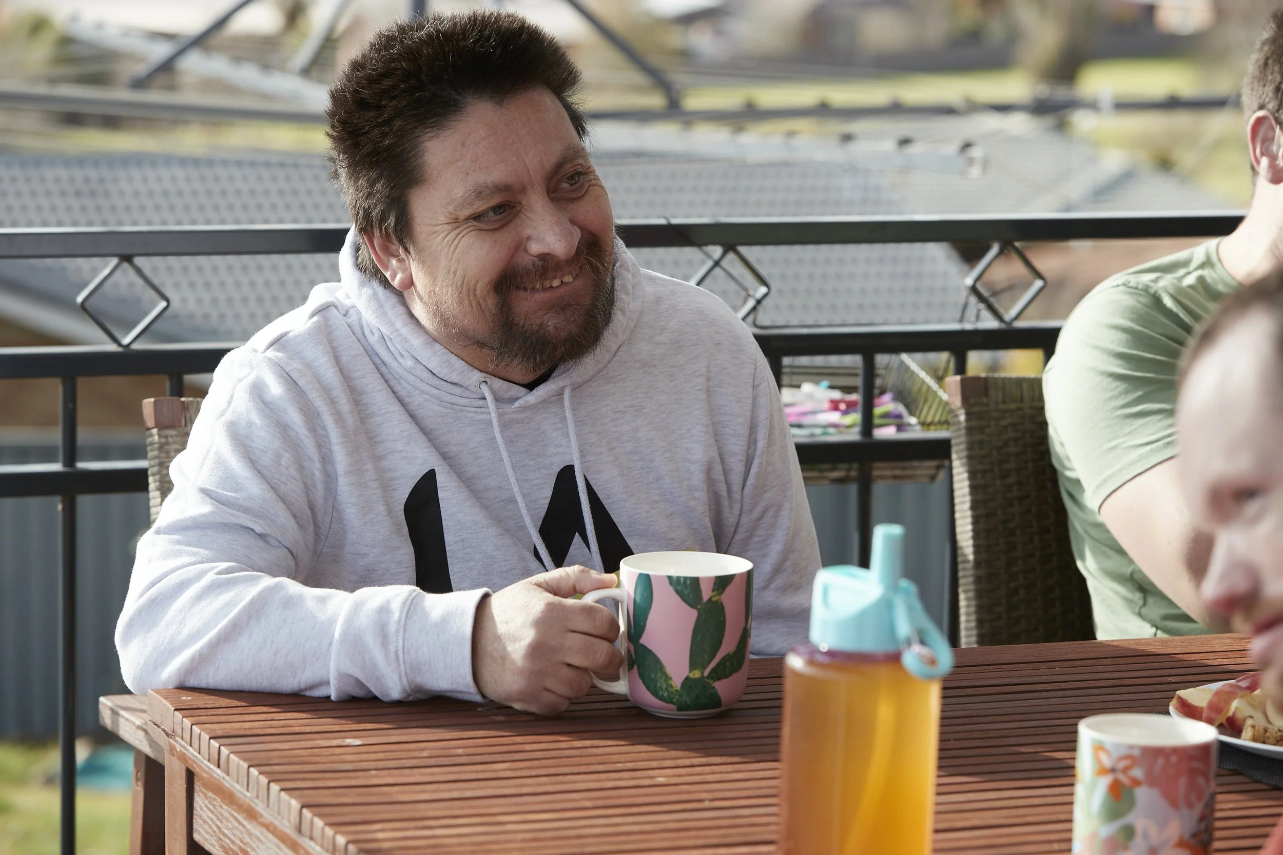 man sitting at a wooden table with a mug and smiling. He is outside on a deck.