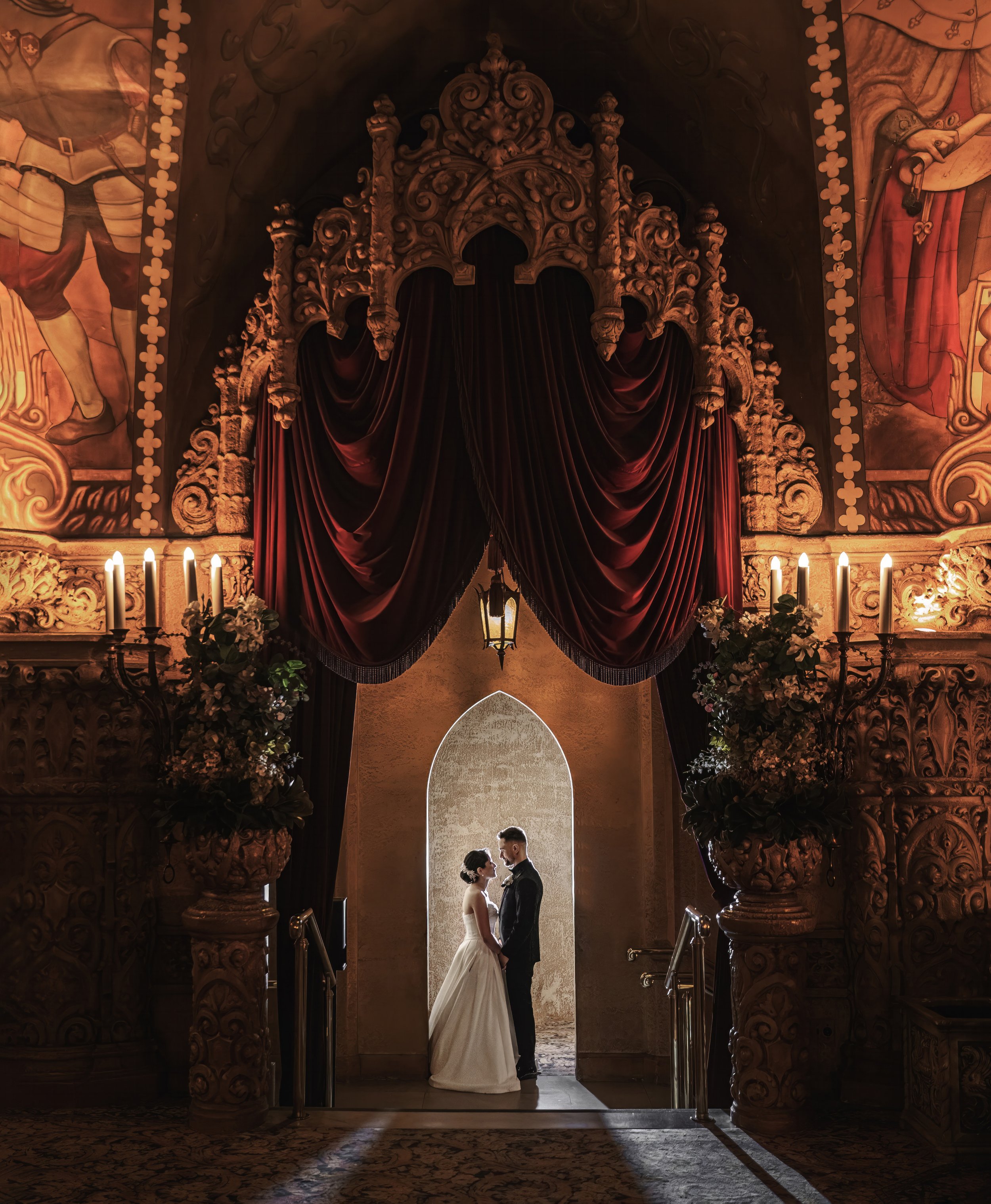 A mesmerizing photo set inside the Regent Theatre in Melbourne, capturing the bride and groom standing under a doorway, holding both hands and staring deeply into each other's eyes. The opulent surroundings of the theatre, with its elaborate architec