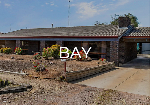 Single-story brick house with a sloped roof, front yard with shrubs and flowers, concrete driveway, and a sign that reads 'BAY'.