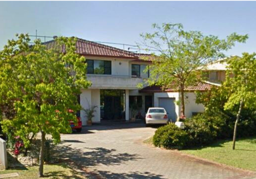 Residential house with two stories, white exterior, red tile roof, surrounded by trees and shrubs, with a driveway and parked cars.