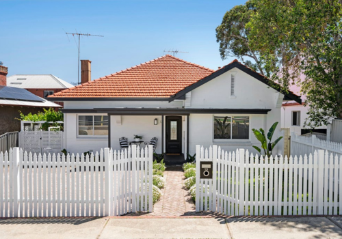 Front view of a white house with a red-tiled roof, black front door, and white picket fence, situated in a suburban neighborhood.