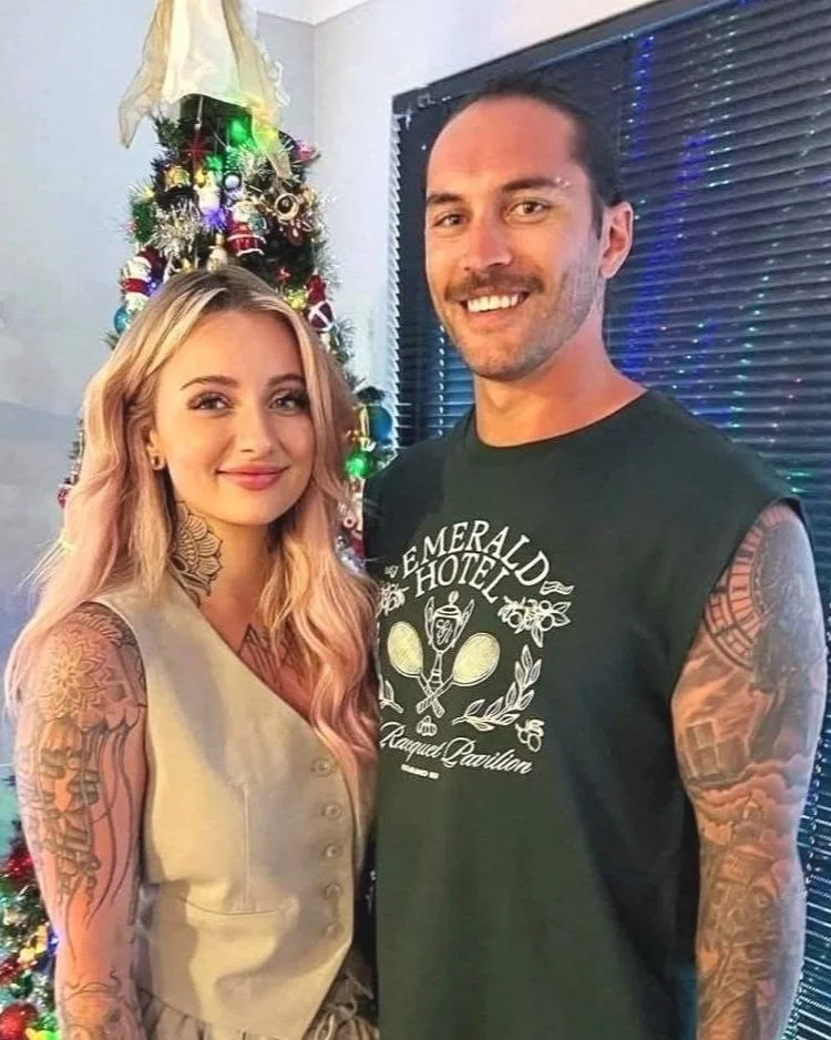 A woman with long blond hair and tattoos on her arms standing next to a man with short dark hair, a beard, and tattoos on his arms, in front of a decorated Christmas tree and a window with blinds, both smiling at the camera.