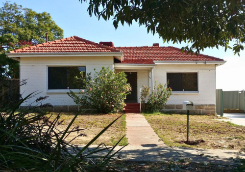 Front view of a single-story house with white brick exterior, red tiled roof, large front windows, and a small front porch with steps, surrounded by a lawn with bushes and a tree, under a bright blue sky.