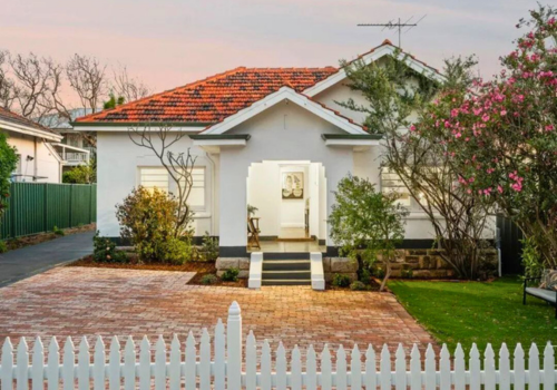 White house with red-tiled roof, front porch, small front yard with plants and blooming trees, brick pathway, white picket fence