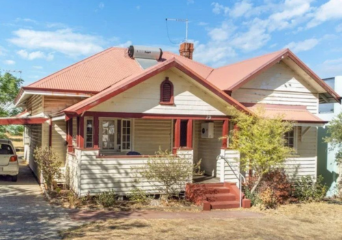 Old two-story house with a red roof, white weathered siding, and a front porch with steps. There are trees and bushes in the yard, and a car is parked on the driveway on the left side.