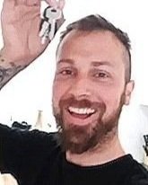 A smiling man with a beard and short hair taking a selfie indoors.