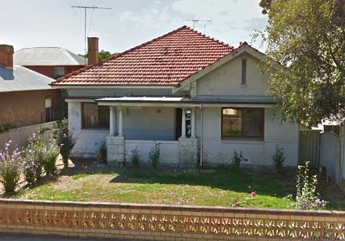 A small, single-story house with a red tiled roof, white walls, and a small front porch. There is a grassy yard with some plants and flowers, and a wooden fence in front.