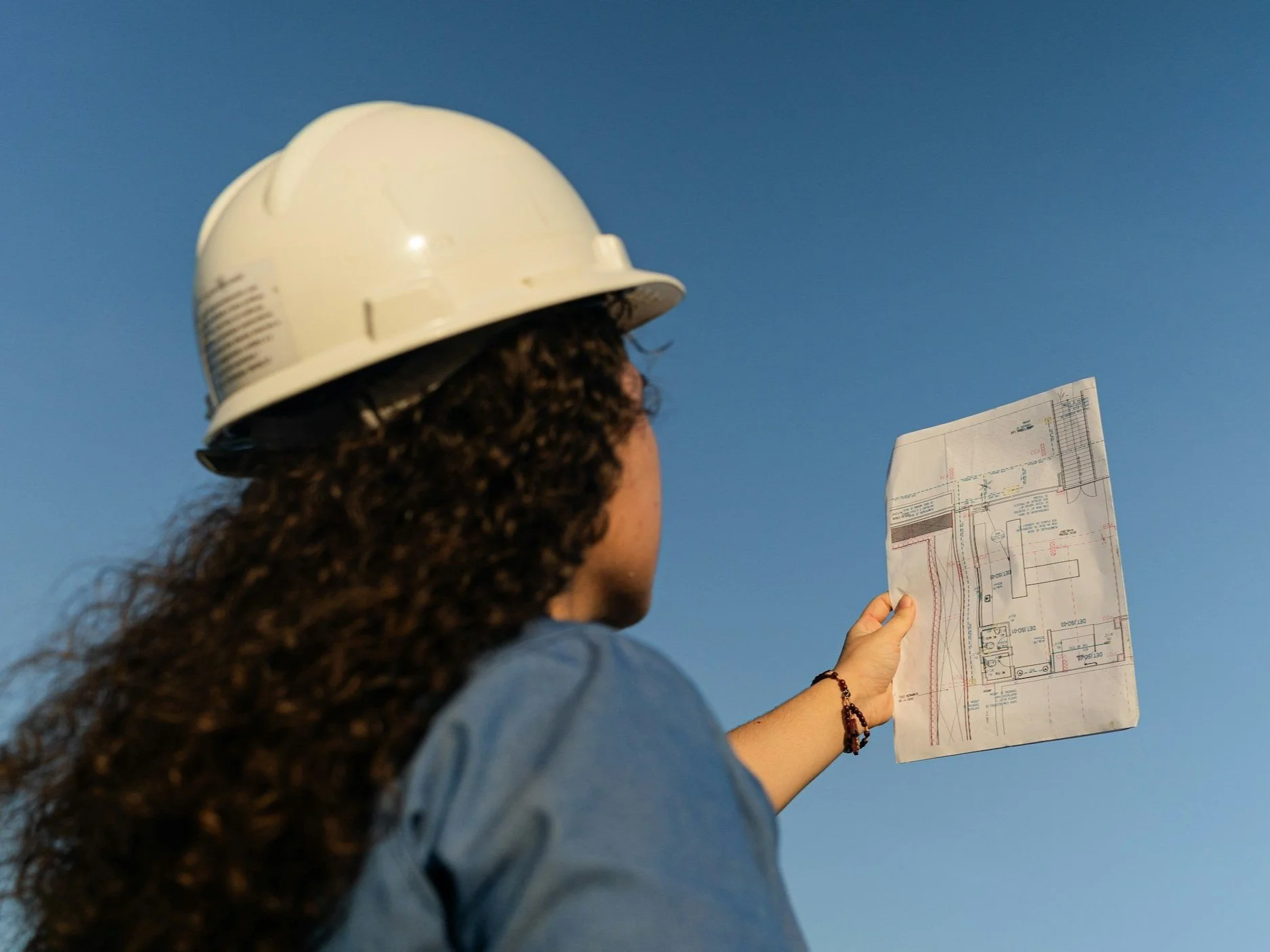 A woman wearing a white safety helmet holding a blueprint against a clear blue sky.