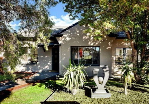 A modern house with large windows, surrounded by trees and greenery, featuring a decorative sculpture in the front yard.