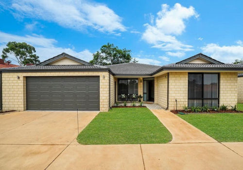 A single-story house with a brick exterior, a two-car garage, a small front porch with a black door, and well-maintained lawn and sidewalk under a blue sky with some clouds.