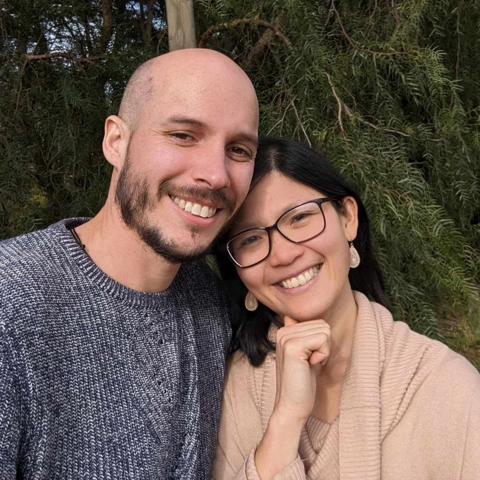 A smiling man with a beard and a woman with glasses, both posing together outdoors in front of trees.