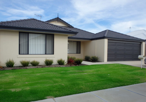 A modern single-story house with a dark gray tiled roof, beige exterior walls, and a two-car garage painted in dark gray. The front yard has well-maintained green grass and small landscaped bushes.