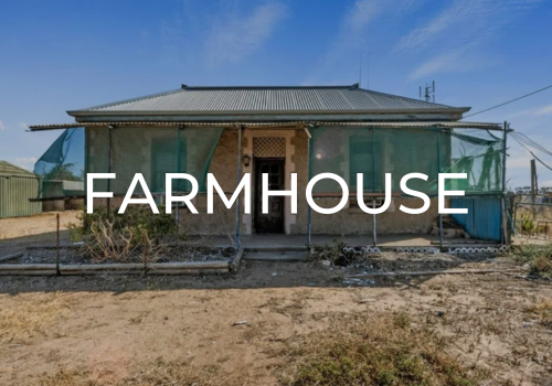 A small, rustic farmhouse with a metal roof, surrounded by a dirt yard and green protective netting.