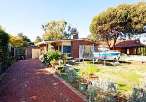 A mid-century modern brick house with a curved roof, surrounded by a garden with potted plants, a brick driveway, and a boat on a trailer in the yard, under a large tree and clear blue sky.