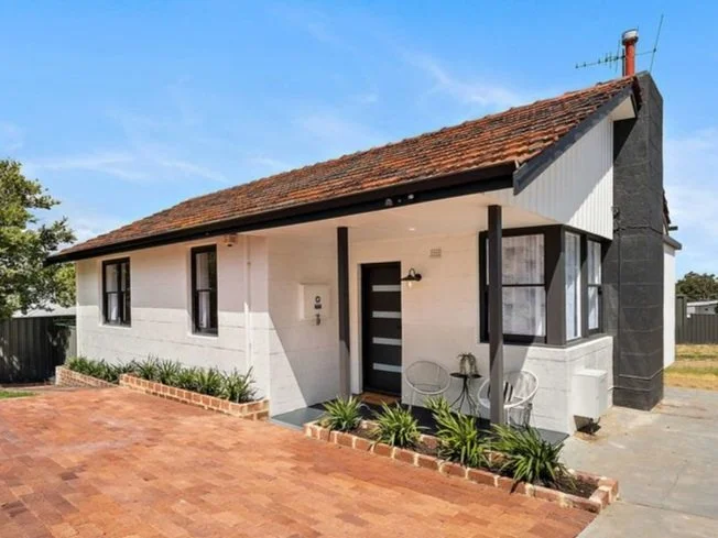 A small white house with a tiled roof, black window frames, a black front door, and a small porch with outdoor seating and garden plants in front.