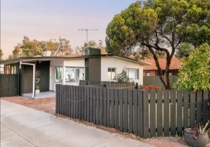 A single-story house with a dark gray fence and a large tree in front, with some shrubs and a sidewalk.