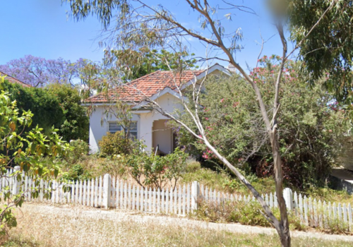 A white house with a red shingle roof surrounded by trees and a white picket fence under a blue sky.