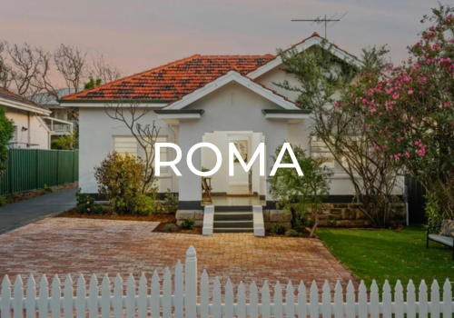 A white house with a red-tiled roof, front steps, and a small front yard with trees, bushes, and a white picket fence, with the text 'ROMA' overlaid.