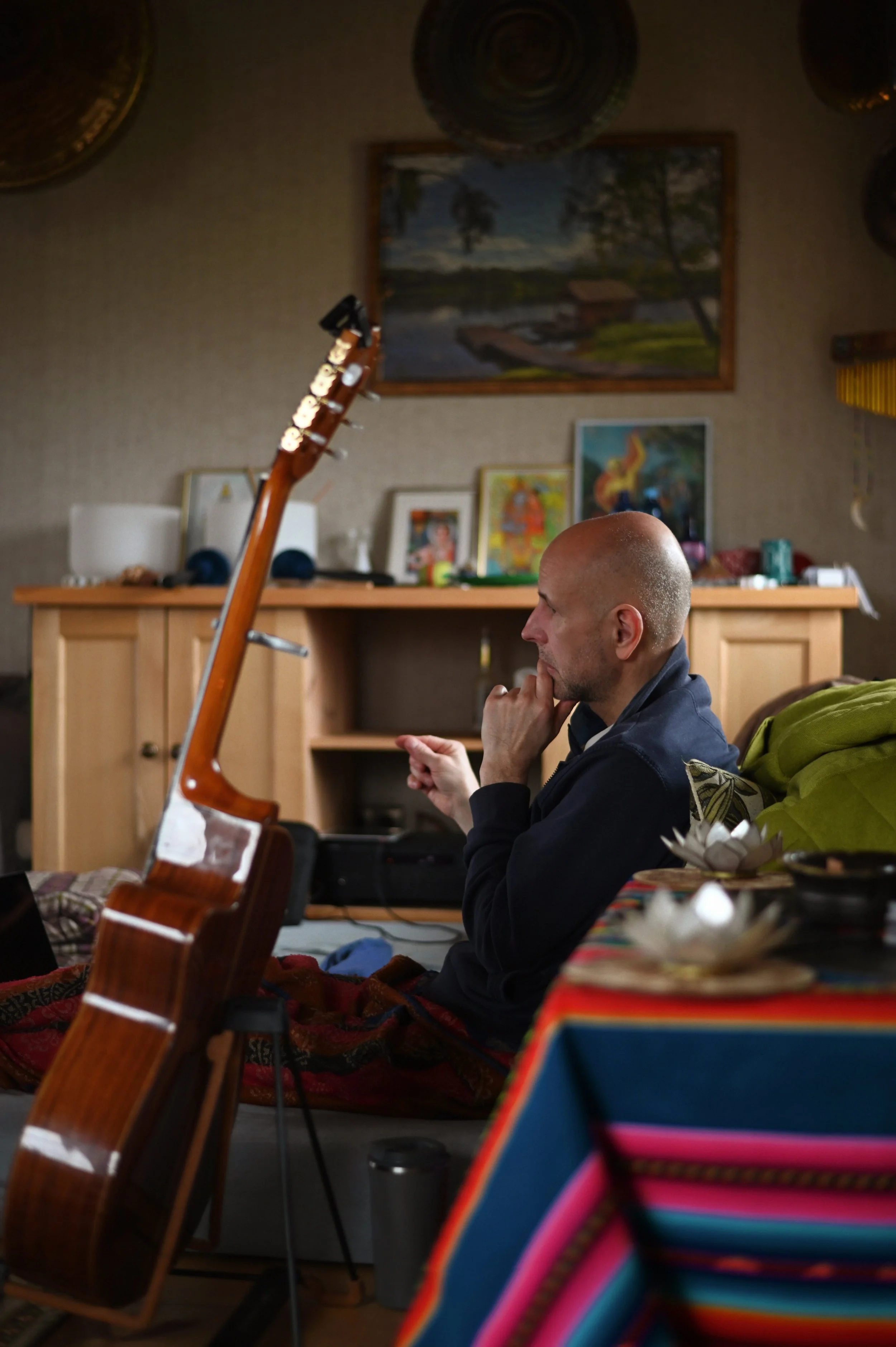 A man sitting on a couch with a guitar in front of him, resting his chin on his hand, in a living room with artwork and decorative items.