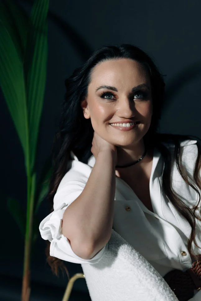 A woman with long dark hair and makeup, smiling and resting her chin on her hand, sitting on a white chair with a green plant in the background.