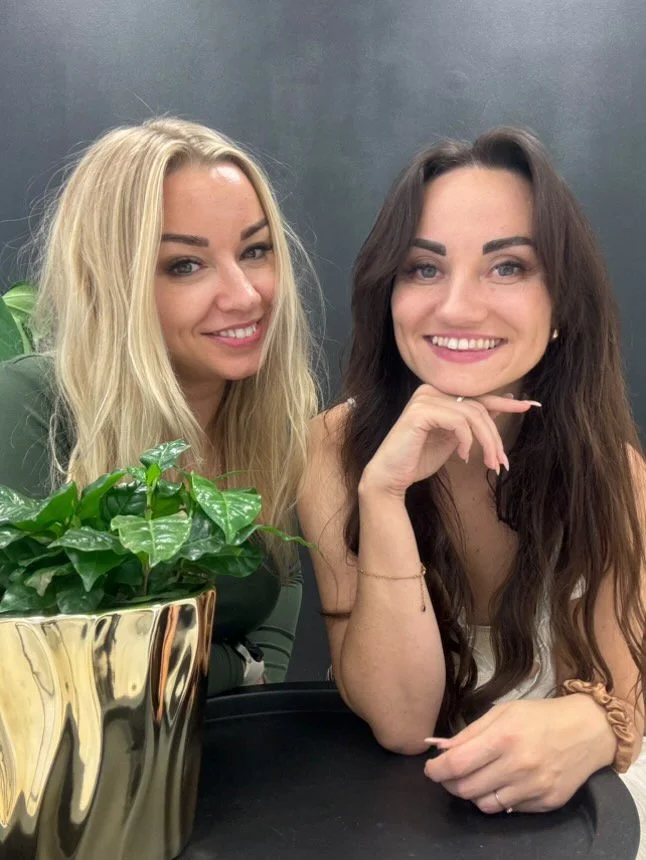 Two women sitting at a table with a potted plant, smiling at the camera, against a gray background.