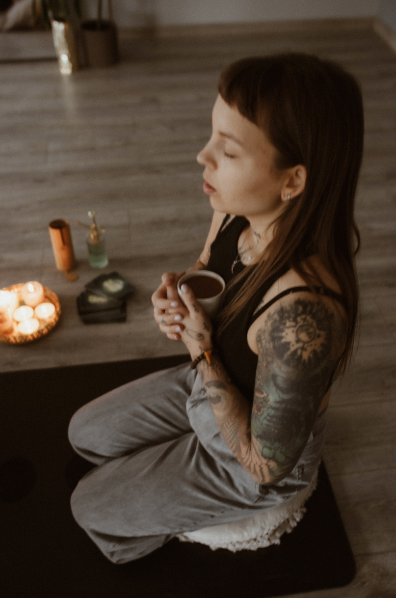 A person with tattoos is sitting on a cushion holding a cup, surrounded by lit candles and a small bottle on a wooden floor.