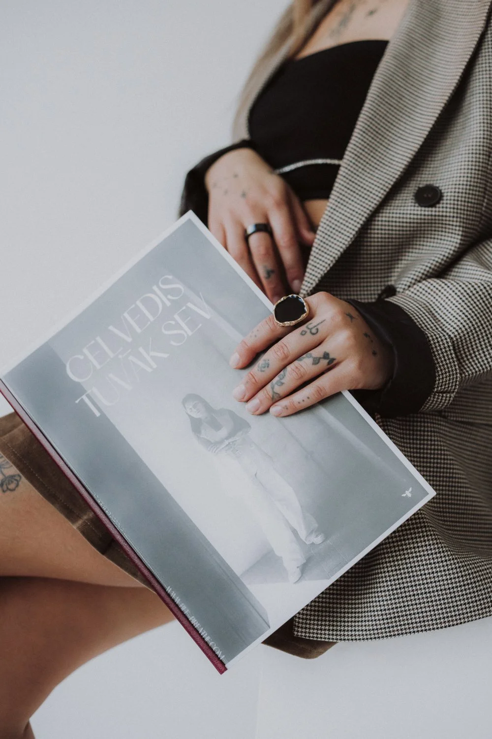 Person holding a book titled 'Celtic Vikings' with tattoos and rings, wearing a houndstooth blazer and black top, sitting with their hand on their stomach.