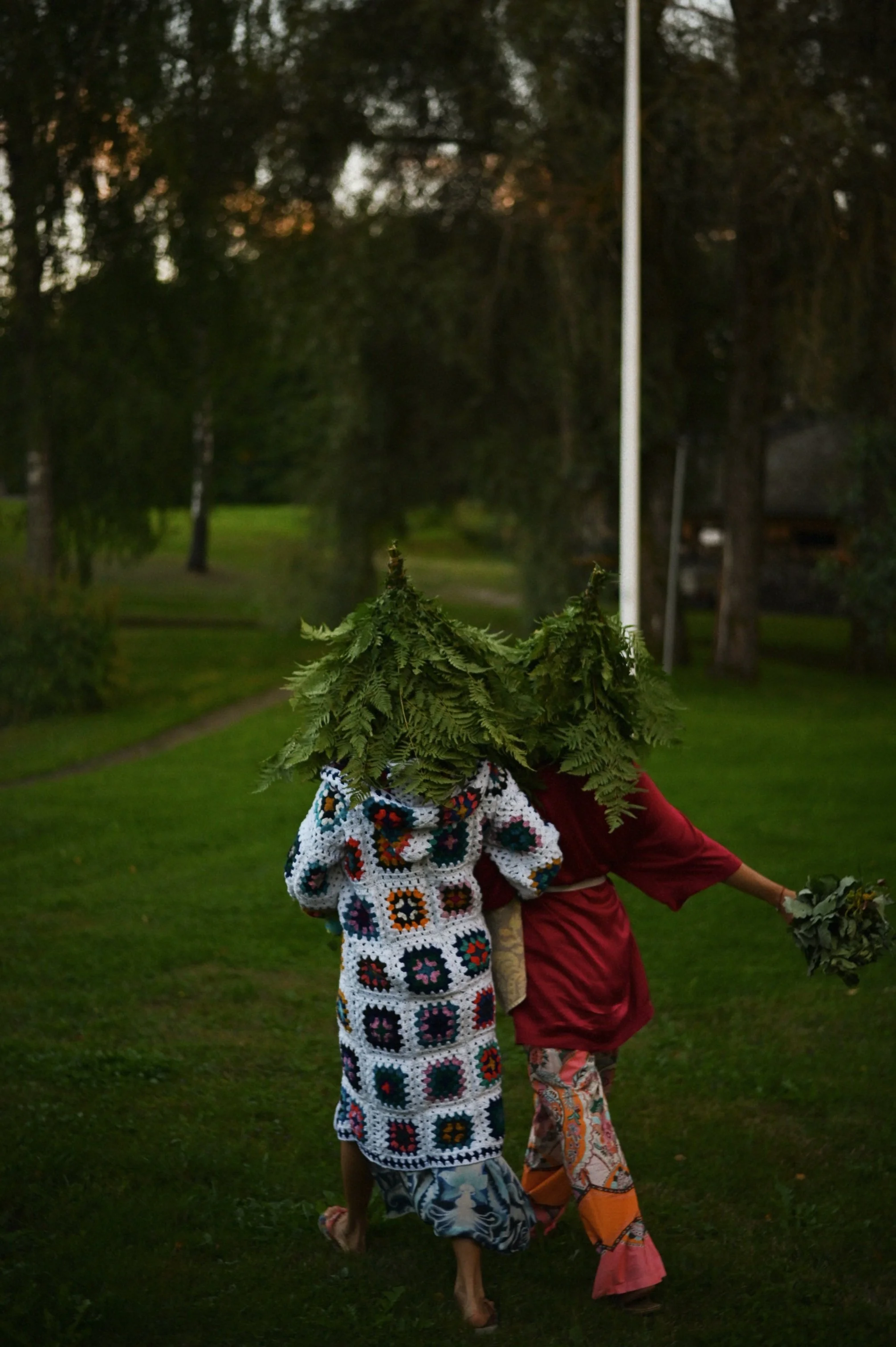 Two people walking away with tree branches on their heads, holding flowers, in a park at dusk.