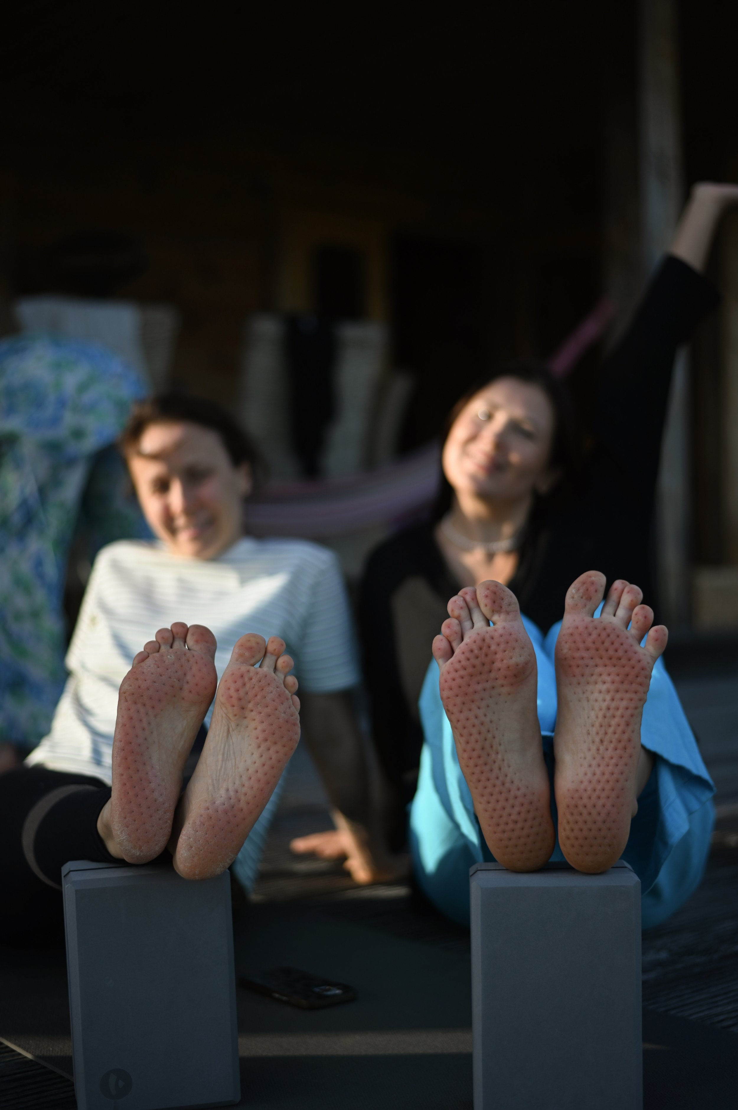 Two women with their feet up in the foreground, showing textured soles, while sitting on a wooden deck outside with a blurred background of chairs and a hammock.