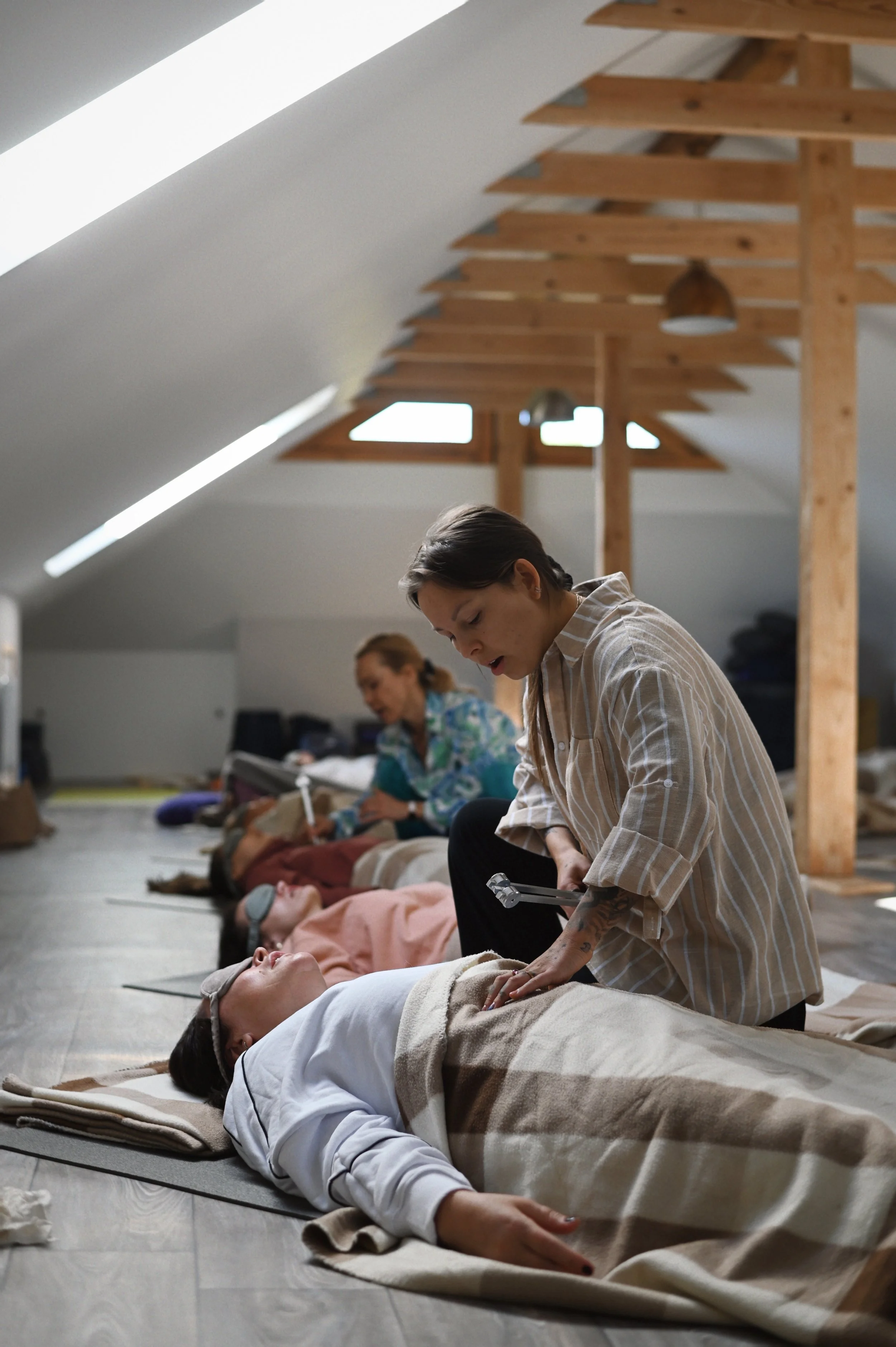 A group of people lying on the floor, receiving guided meditation or relaxation, with an instructor leaning over one participant, in a room with sloped ceiling and wooden beams.