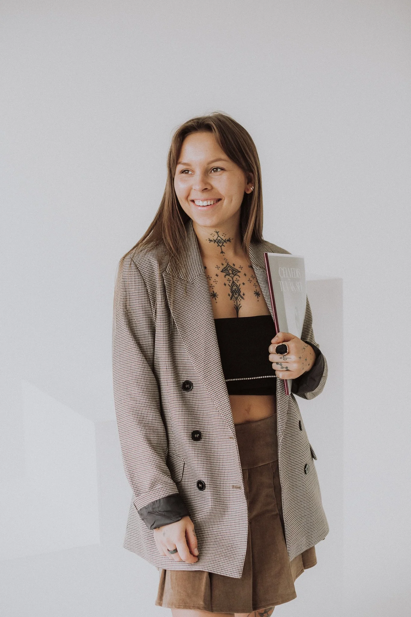Person wearing a blazer and holding a book, with visible tattoos on their neck and hand.