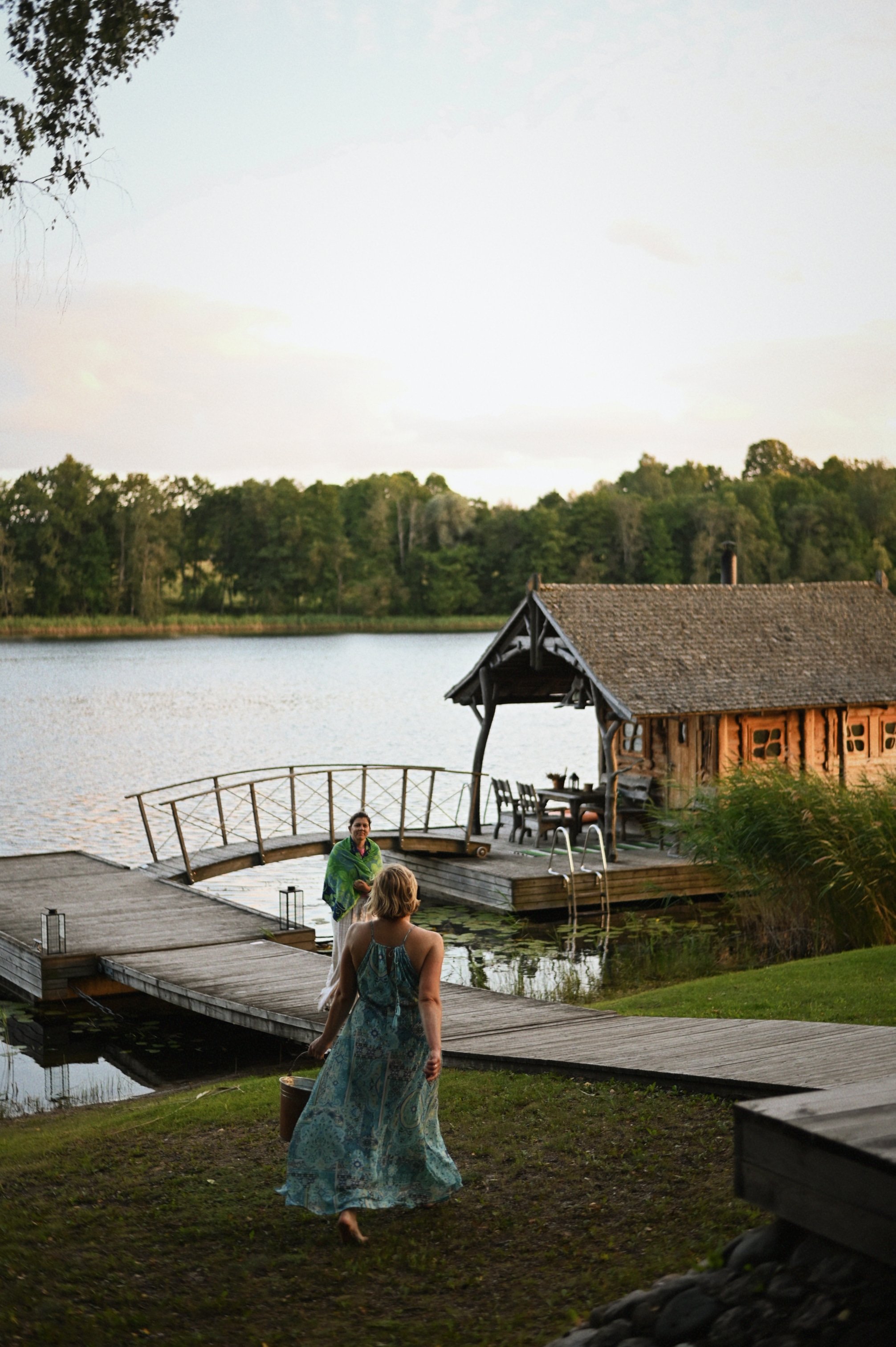Two women are near a pond, one walking into the scene holding a bucket and the other standing on the dock by a small rustic cabin, with trees and water in the background during sunset.