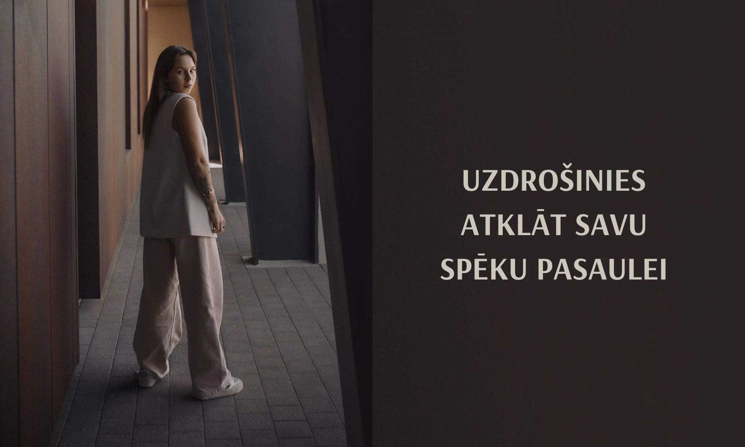 A woman standing in a hallway, looking towards the camera, with wooden panel walls on one side and a row of vertical panels on the other side. The right side of the image contains a dark background with white text in Lithuanian that reads, "Uzdrošinies atklāt savu spēku pasaulei."