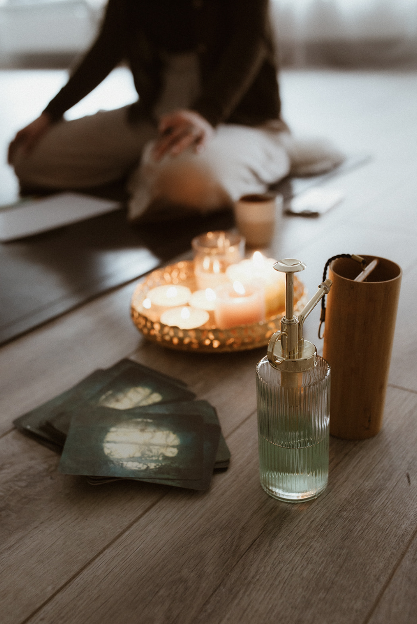 A meditation setup with candles on a tray, tarot cards, and a glass spray bottle on a wooden floor. A person is sitting cross-legged in the background.