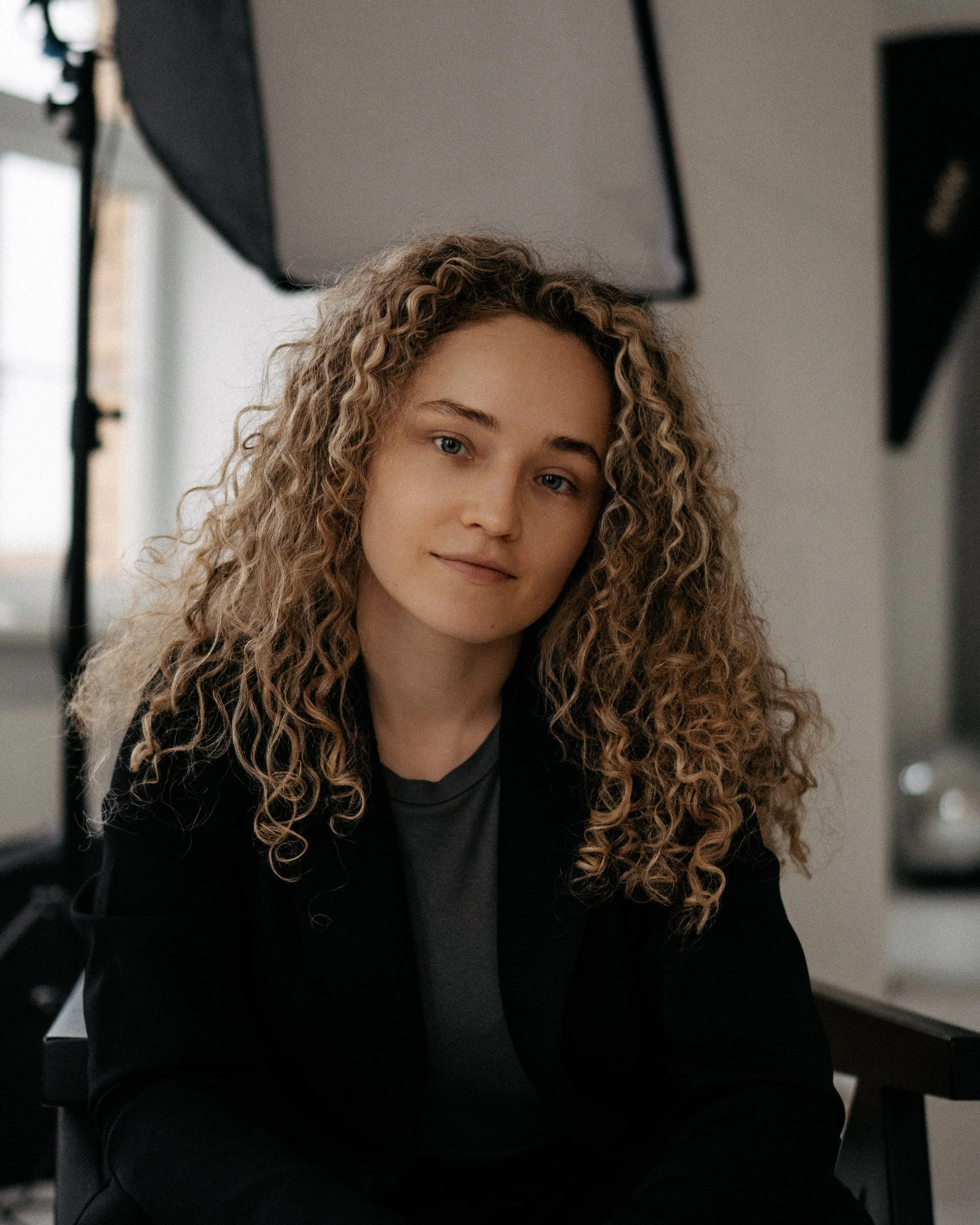 A woman with curly blonde hair, wearing a black blazer over a gray top, sitting in a photography studio with soft lighting and professional equipment in the background.