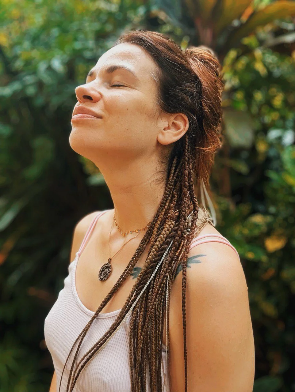 A woman with braided hair, wearing a tank top and jewelry, standing outdoors with eyes closed, smiling peacefully amidst green foliage.