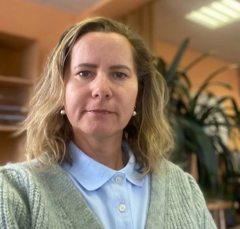 A woman with shoulder-length wavy blonde hair, wearing pearl earrings, a light blue collared shirt, and a light green sweater, standing in a room with wooden shelves and a large plant in the background.