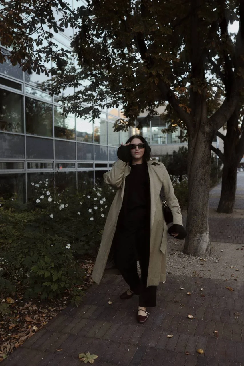 Woman wearing sunglasses, a beige trench coat, black pants, and heels, walking on a brick sidewalk beside a tree and a building with glass windows.