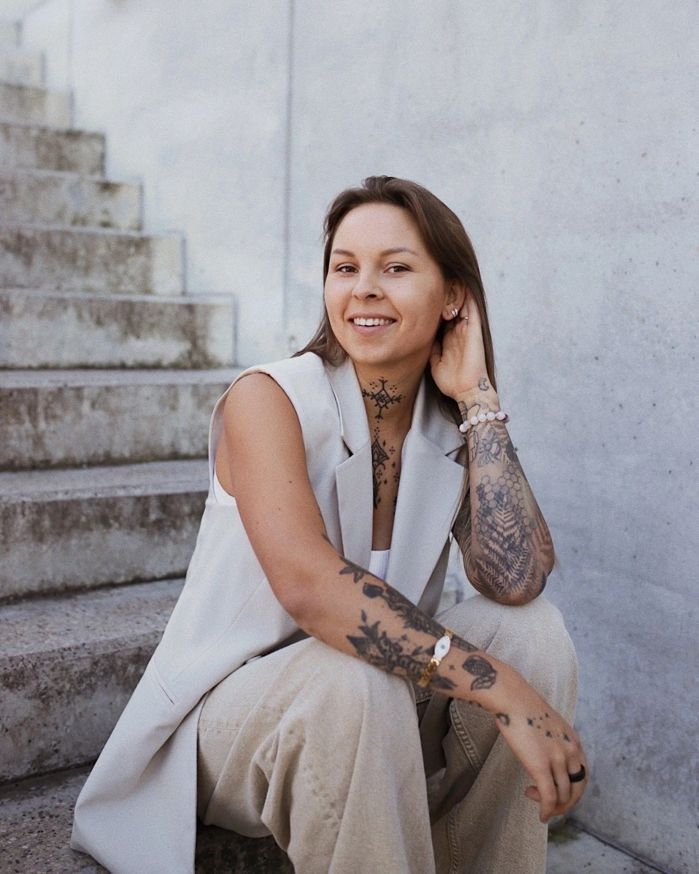 A woman with tattoos on her arms and neck, sitting on stairs, smiling, wearing a sleeveless vest and beige pants, with a gray wall in the background.