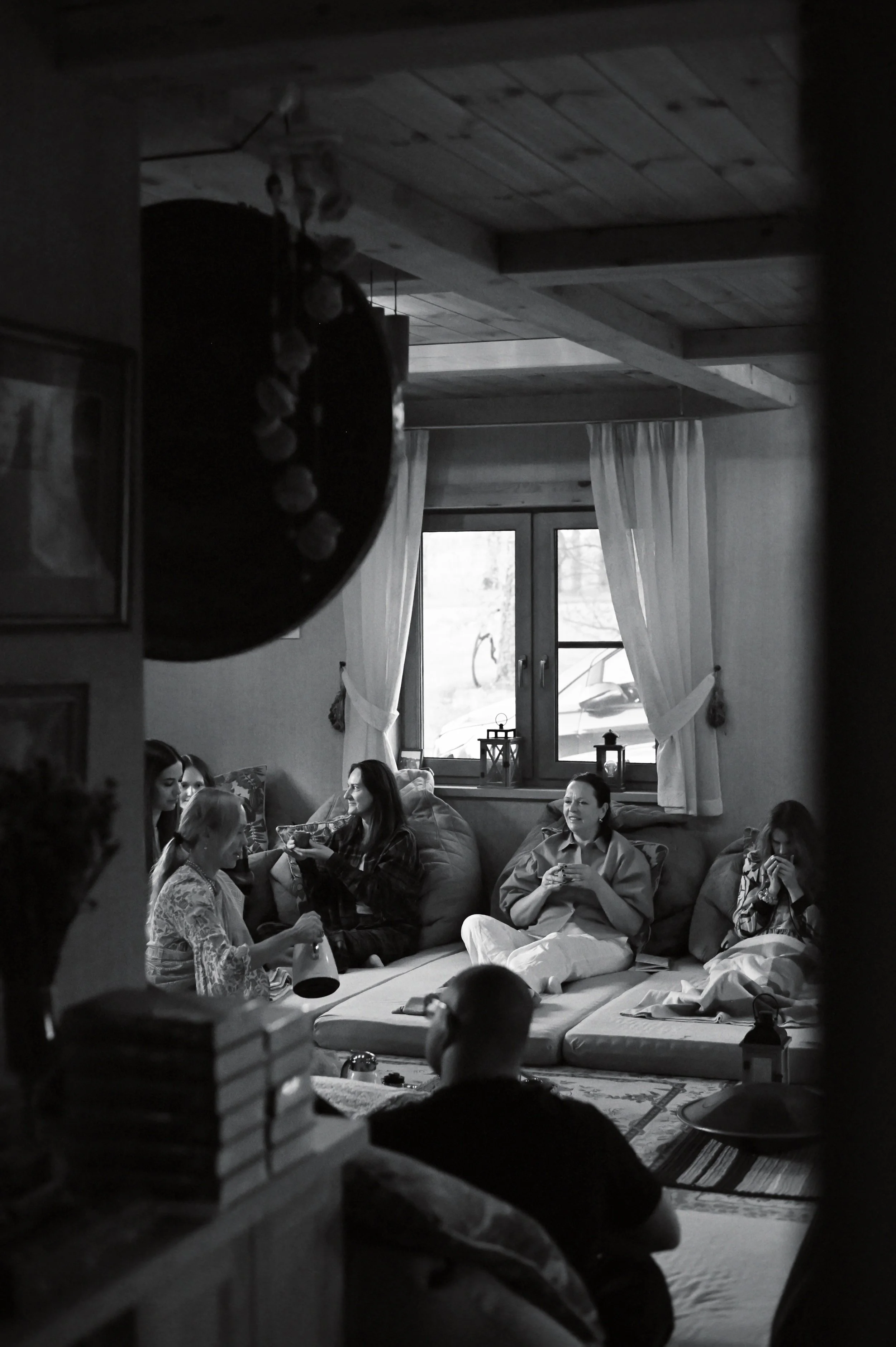A group of women sitting on floor cushions in a cozy living room, some drinking from cups and one pouring a drink, with a large window and curtains in the background.