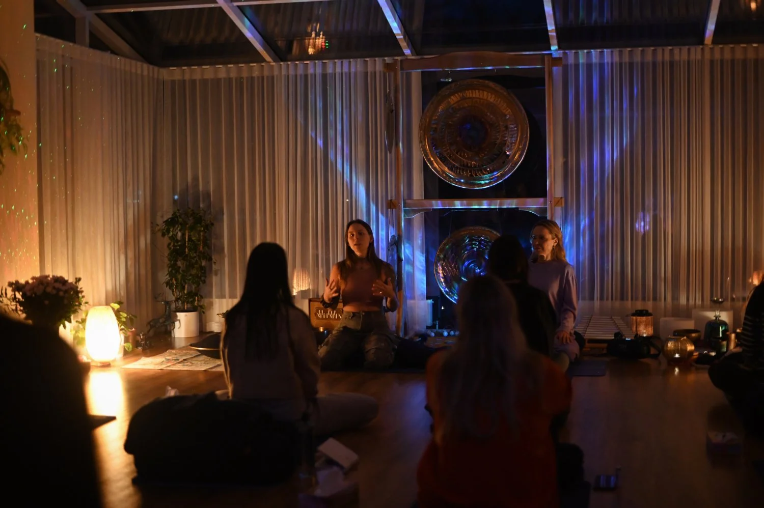 A group of people participating in a meditation or yoga session in a dimly lit room with decorative lighting and large metallic orbs on the wall.