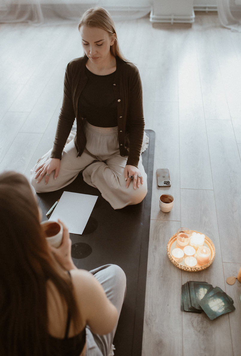 Two people sitting on a floor with a black mat, one facing the camera holding a cup, the other sitting cross-legged with a piece of paper and pen. A phone, candles, and cards are placed on the floor nearby.