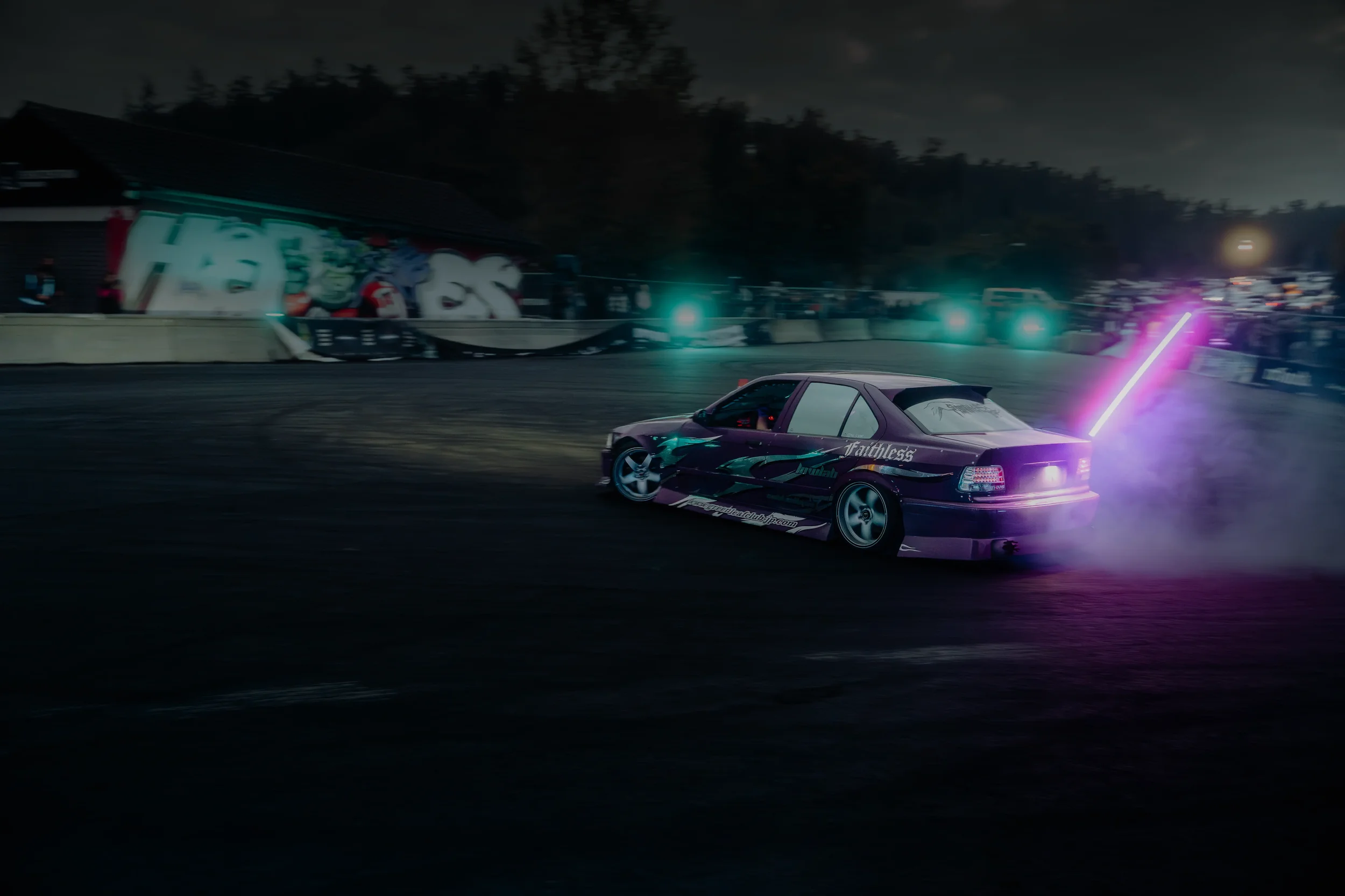A purple racing car with digital graphics and text on the side, emitting pink and purple smoke, drifting on a dark race track at night with colorful lights in the background.