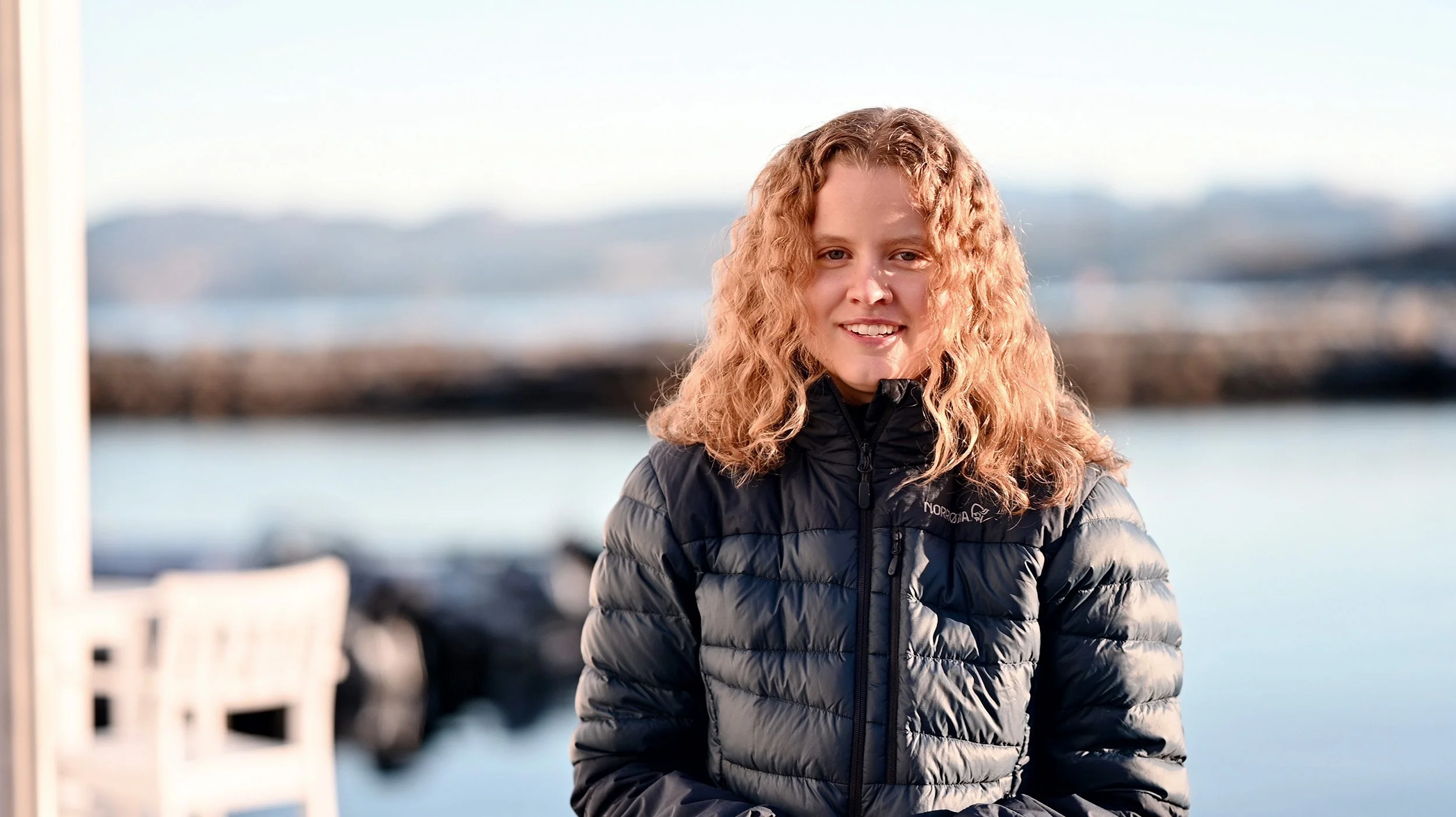 En smilende jente med krøllete, lys håret, iført en svart dunjakke, som står foran vann med en bro og fjell i bakgrunnen.