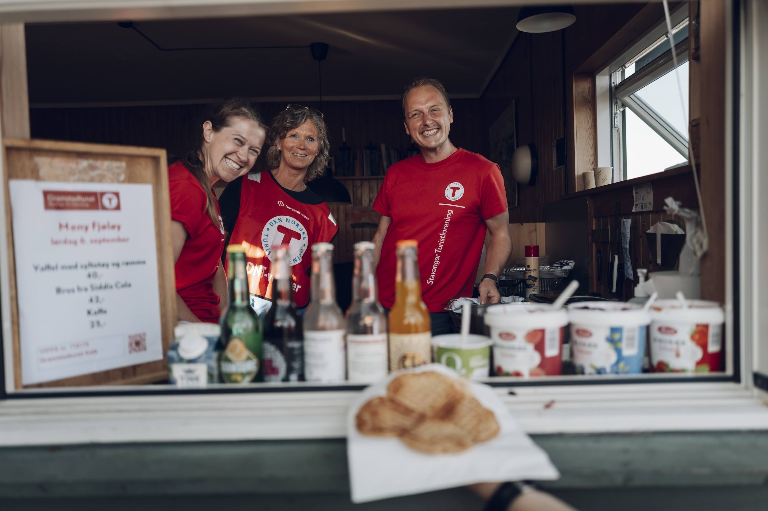 Tre personer bak en kafé-/salgsbod som selger is og drikke. De smiler mot kamera, og er iført røde T-skjorter med logo og tekst.