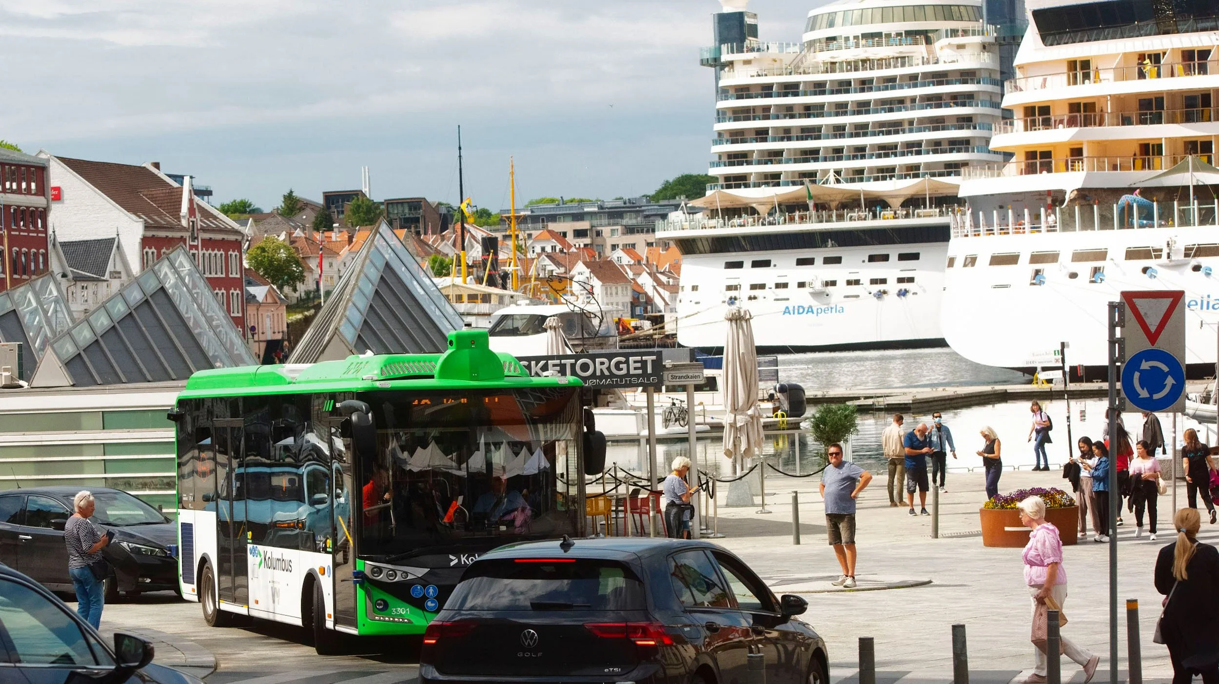 Utsikt over en havn med båter og kryssningsskip, mennesker som går og venter, og en grønn og hvit bus ved en holdeplass.
