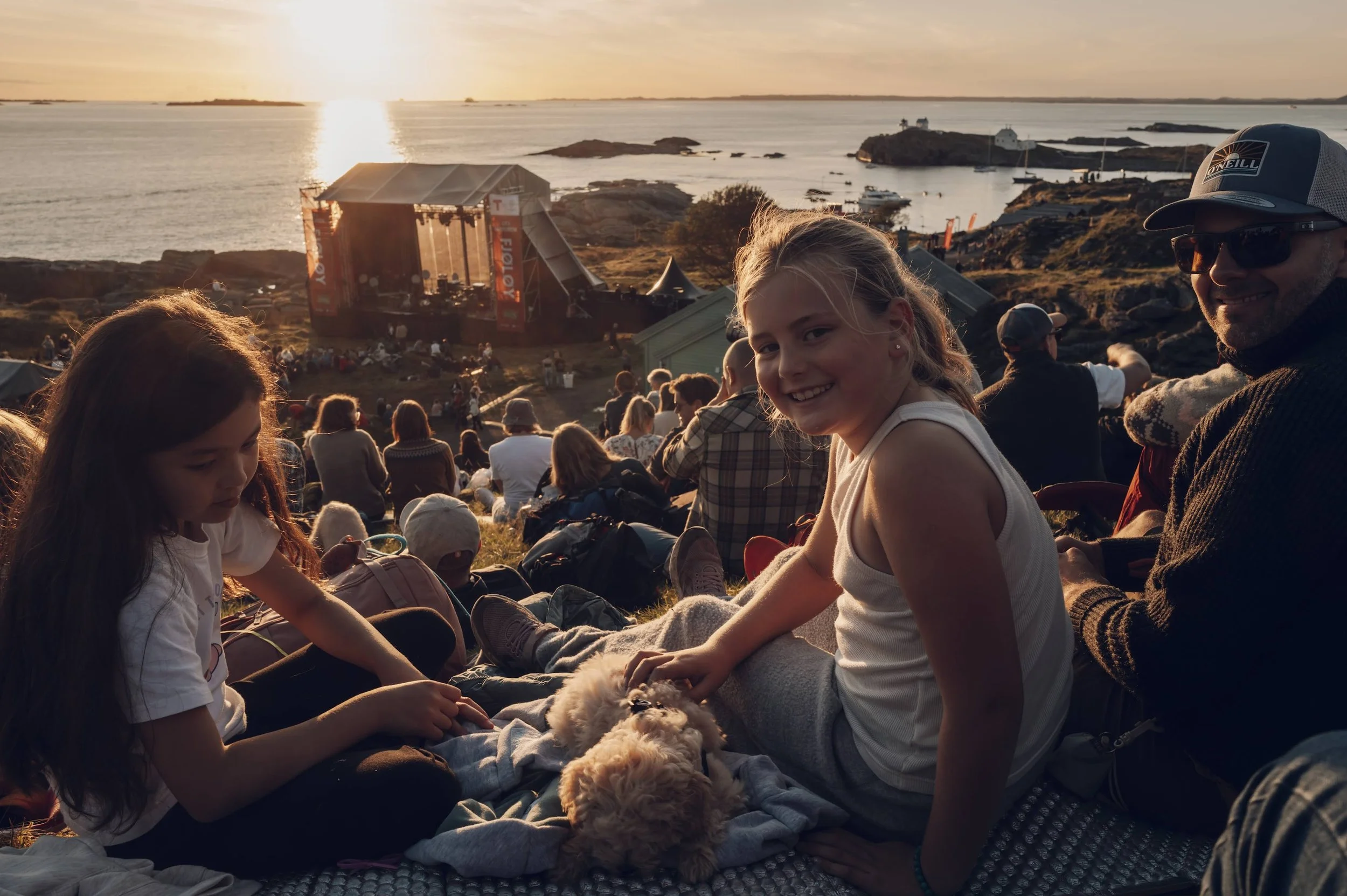 Folk sit på en skråning og ser på en konsert ved solnedgang ved en havn, hvor et utendørs scene er satt opp.