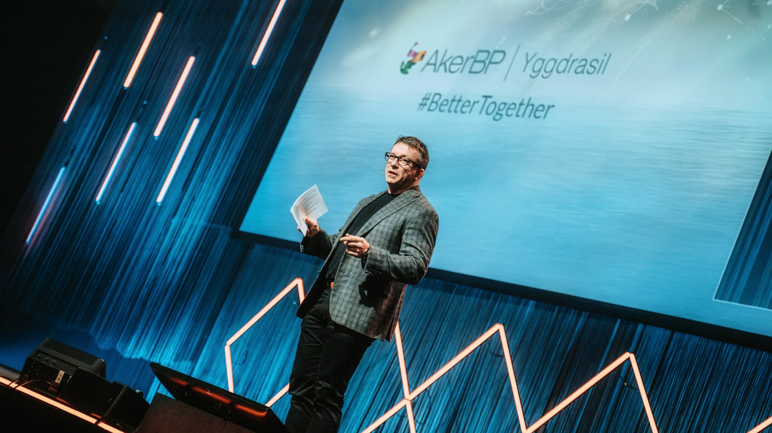 En mann holder en presentasjon på en scene med en stor skjerm bak seg som viser logoer og hashtags, med blå og oransje lys dekorasjoner.