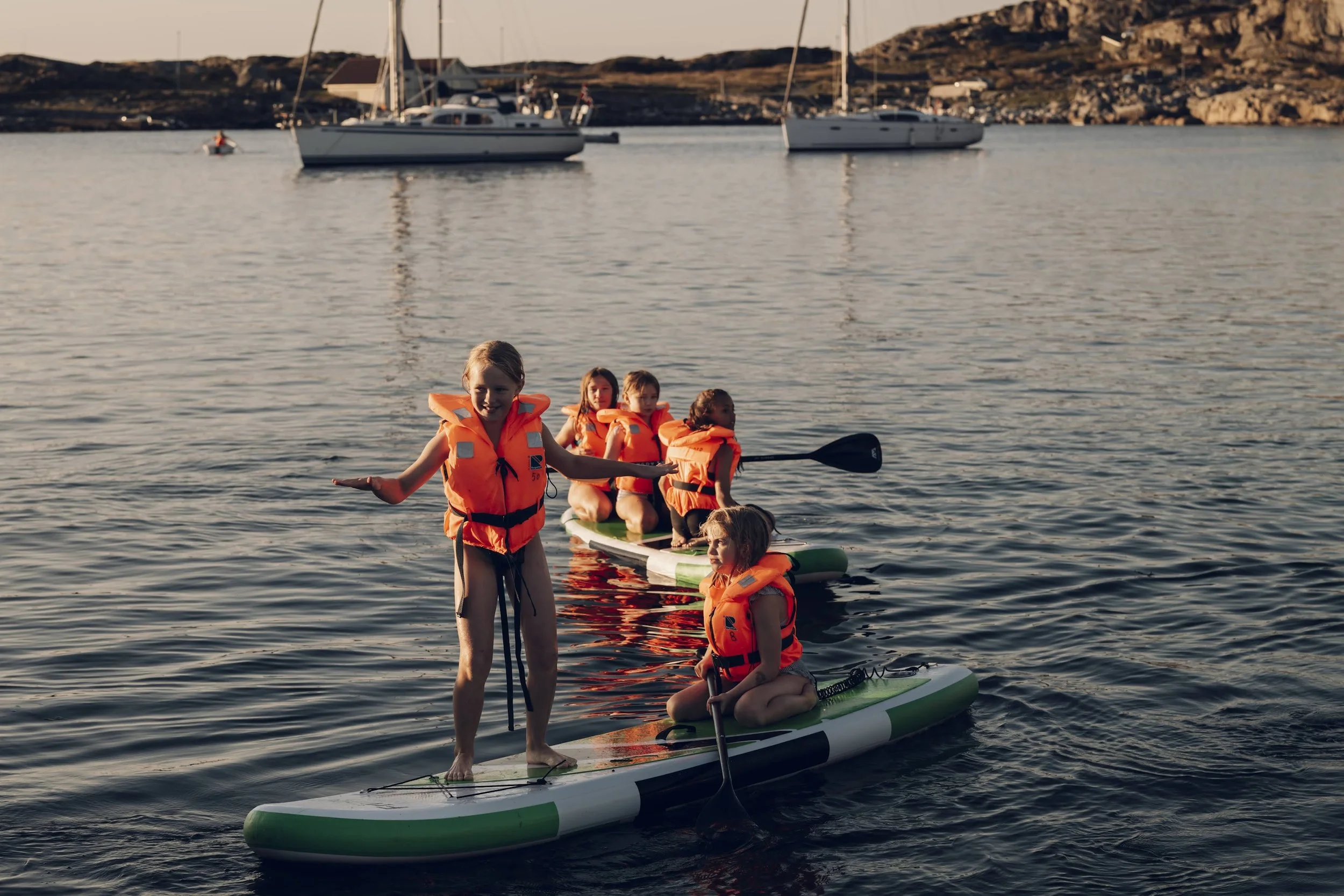 Fem barn på stående og sittende paddleboard i vann, alle iført orange redningsvester, med båter og øy i bakgrunnen i solnedgangen.